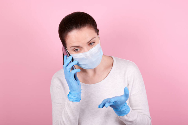 Portrait of a brunette girl in a medical mask and blue rubber gloves speaking on cellphone. Isolated on pink background. - Photo, image