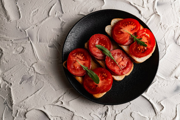 Three sandwiches with fresh and bright red tomatoes and smoked sausage, decorated with a leaf of green arugula on a matte black plate and a white stone background - Foto, immagini