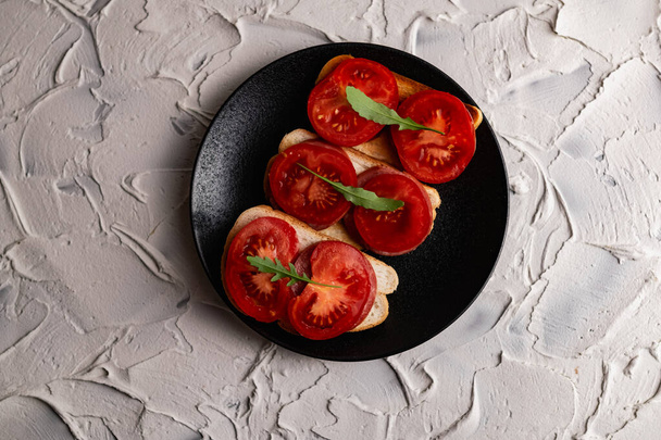 Tre panini con pomodori rossi freschi e brillanti e salsiccia affumicata, decorati con una foglia di rucola verde su un piatto nero opaco e uno sfondo di pietra bianca - Foto, immagini