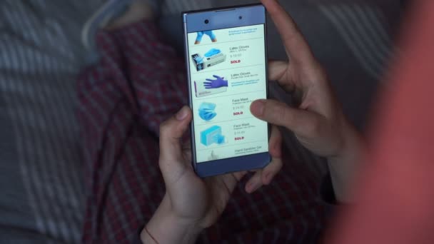 A closeup on a hand holding a smartphone, scrolling through a page. Online shopping for necessities such as toilet paper, hand sanitizer , disinfectant wipes, face masks, latex gloves due to covid-19 - Footage, Video