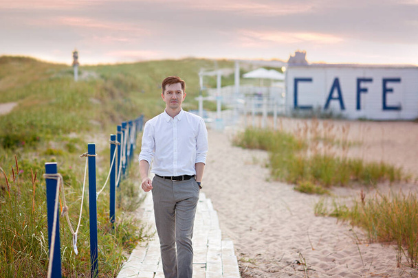 Nuori komea, menestynyt mies valkoisessa paidassa kävelee pitkin puista pakkaa meren rannalla. Taustalla kaiverrus Cafe - Valokuva, kuva