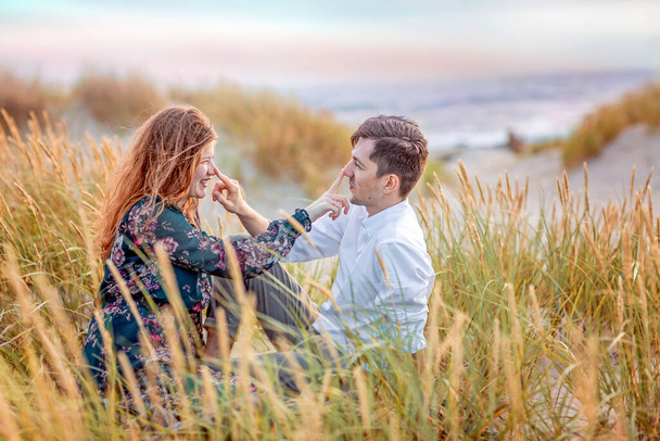 Una giovane coppia innamorata siede tra le dune ricoperte di erba alta. Si toccano scherzosamente il naso a vicenda. Un giovane con una camicia bianca, una ragazza con un vestito verde. - Foto, immagini