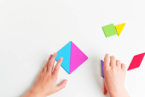 Tangram barevné geometrické puzzle kousky s childs ruce pohybující se kusy na bílém stole, horní pohled.  - Fotografie, Obrázek