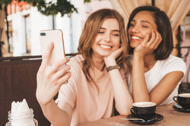 Two young beautiful smiling hipster girls in trendy summer clothes.Carefree women chatting in veranda cafe and drinking coffee.Positive models having fun and and taking selfie on smartphone - Φωτογραφία, εικόνα