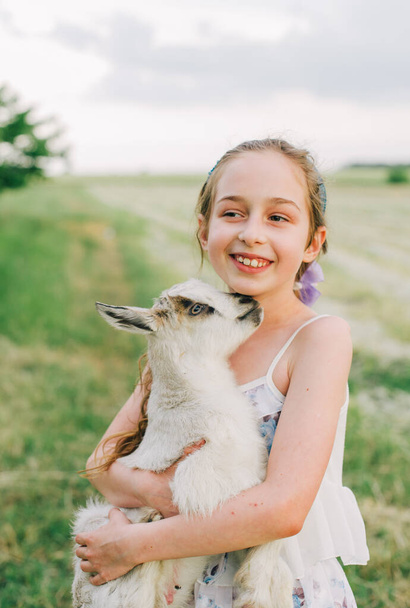 girl with baby goat on farm outdoors. Love and care. Village animals. happy child hugs goat, concept of unity of nature and man. Friendship of child and animals. Happy childhood. Girl and goat - Photo, image
