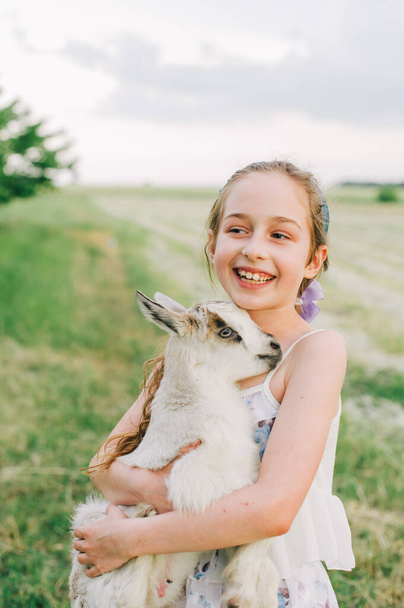 dívka s kozou na farmě venku. Lásku a péči. Vesnická zvířata. šťastné dítě objímá kozu, pojetí jednoty přírody a člověka. Přátelství dětí a zvířat. Šťastné dětství. Dívka a koza - Fotografie, Obrázek
