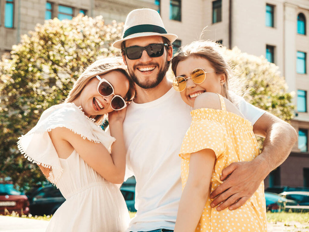 Group of young three stylish friends posing in the street. Fashion man and two cute girls dressed in casual summer clothes. Smiling models having fun in sunglasses.Cheerful women and guy going crazy - Фото, изображение