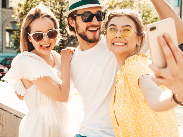 Group of young three stylish friends in the street.Man and two cute girls dressed in casual summer clothes.Smiling models having fun in sunglasses.Women and guy making photo selfie on smartphone - Foto, Bild