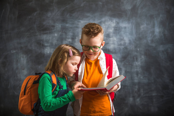 Dwójka dzieciaków bawi się czytając książkę przeciwko tablicy. Chłopiec i dziewczynka ze szkolnymi plecakami. - Zdjęcie, obraz
