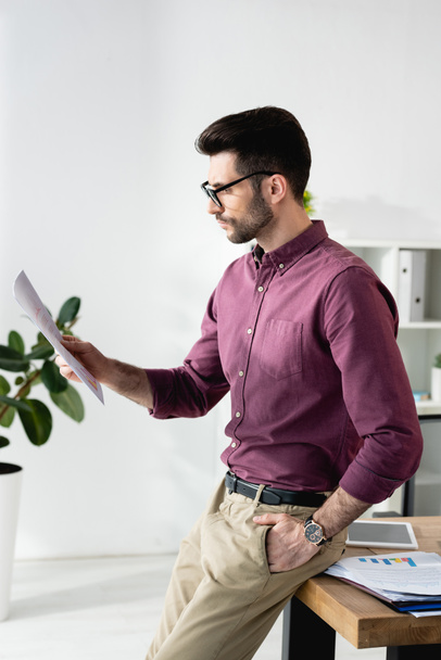 young, serious businessman reading document while leaning on desk with hand in pocket - Foto, Imagen
