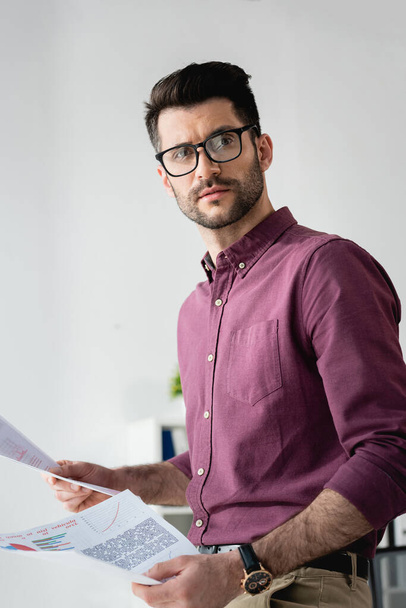 young, serious businessman in eyeglasses looking away while holding documents - Photo, Image