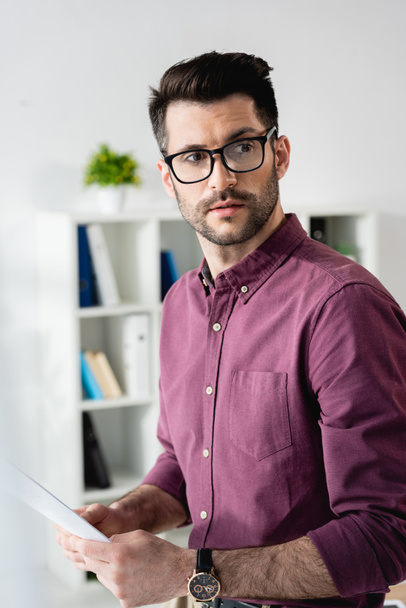 selective focus of serious, handsome businessman in eyeglasses looking away while holding document - Photo, Image