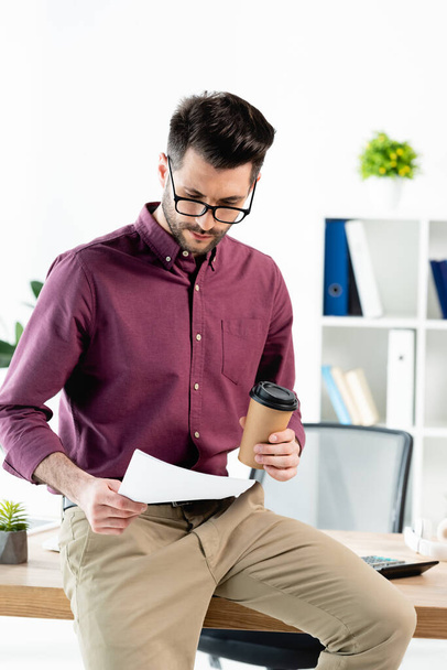 young serious businessman sitting on desk, reading document and holding coffee to go - Foto, immagini
