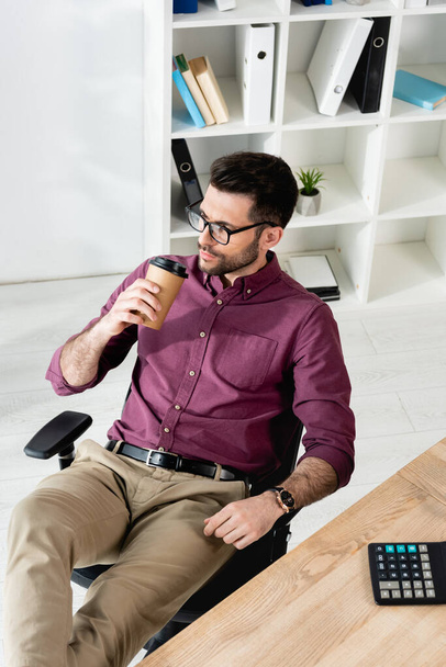 vista ad alto angolo di uomo d'affari sognante bere caffè per andare mentre seduto sul posto di lavoro - Foto, immagini