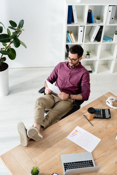 νεαρός επιχειρηματίας ανάγνωση έγγραφο, ενώ κάθεται με τα πόδια στο γραφείο κοντά στο laptop, αριθμομηχανή και καφέ για να πάει - Φωτογραφία, εικόνα