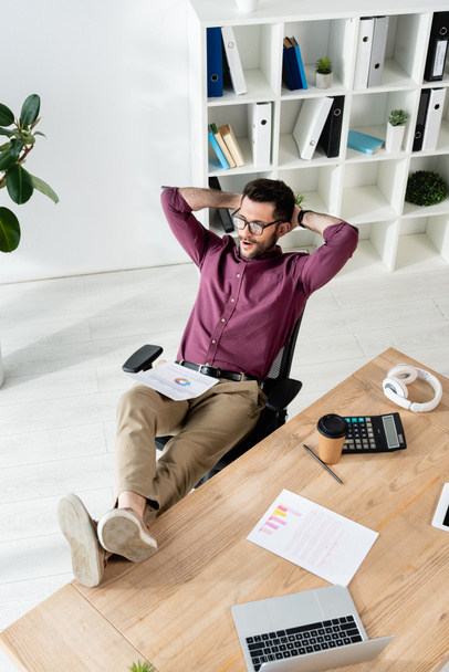 υψηλή γωνία του επιχειρηματία κάθεται με τα πόδια στο γραφείο και τα χέρια πίσω από το κεφάλι κοντά gadgets, χαρτιά και κύπελλο μιας χρήσης - Φωτογραφία, εικόνα