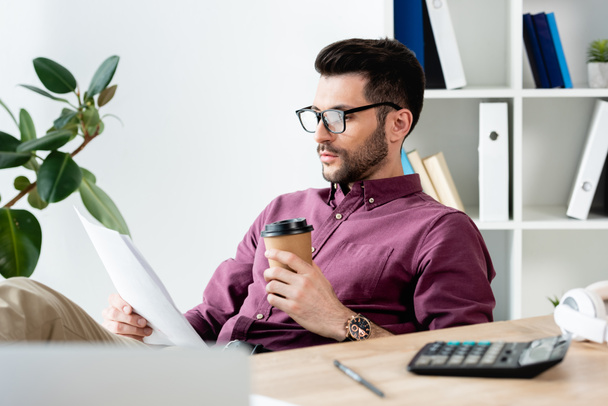 enfoque selectivo del joven empresario sosteniendo una taza de papel mientras lee el documento en el lugar de trabajo
 - Foto, imagen