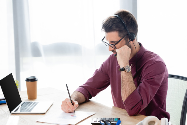 soustředěný podnikatel ve sluchátkách psaní na papíře během online konference na notebooku - Fotografie, Obrázek