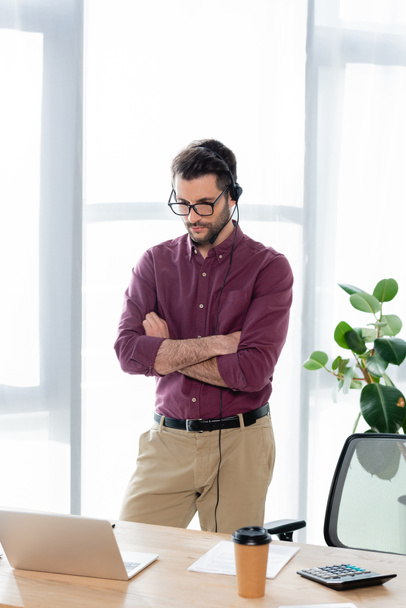 mladý, seriózní podnikatel ve sluchátkách stojící se zkříženýma rukama během video chatu na notebooku - Fotografie, Obrázek