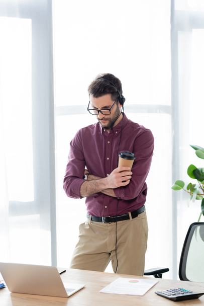 молодой, серьезный бизнесмен в наушниках держит кофе, чтобы пойти во время онлайн-встречи на ноутбуке
 - Фото, изображение