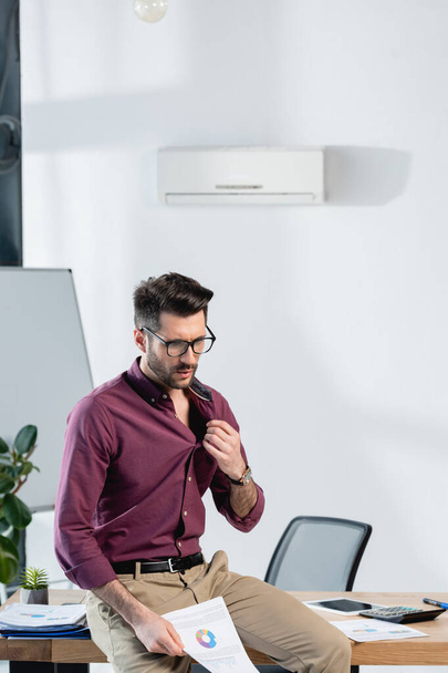 exhausted businessman suffering from heat in office, sitting on desk, touching shirt and holding document - Fotografie, Obrázek