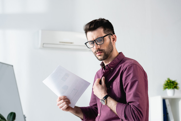 displeased businessman waving paper and touching shirt while suffering from heat in office - Фото, зображення