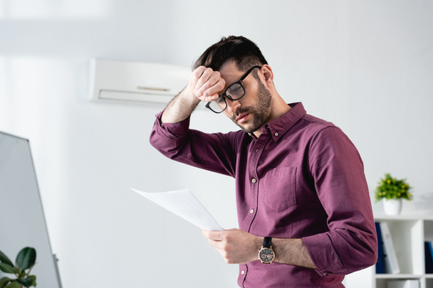 young businessman looking at document and touching forehead while suffering from heat in office - Photo, Image