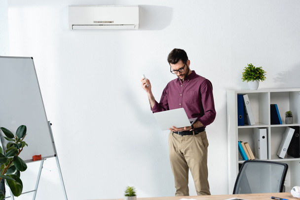 pohledný podnikatel držící notebook a zapínající klimatizaci s dálkovým ovladačem - Fotografie, Obrázek