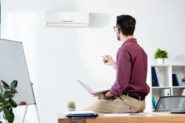businessman sitting on desk with laptop and switching on air conditioner with remote controller - Photo, Image
