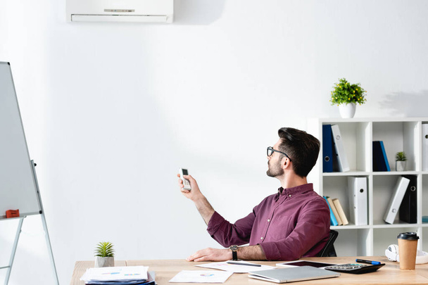 Jungunternehmer sitzt am Arbeitsplatz und schaltet Klimaanlage mit Fernbedienung ein - Foto, Bild
