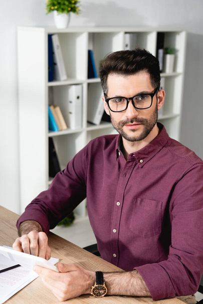 young, confident businessman in eyeglasses looking at camera while using digital tablet - Foto, Imagen