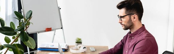 orientation panoramique du jeune homme d'affaires sérieux assis sur le lieu de travail près de tableau à feuilles mobiles et usine verte - Photo, image
