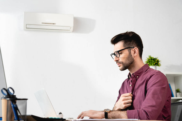 若いビジネスマンがノートパソコンで働いてエアコンの近くで熱に苦しんでいる間にシャツに触れると - 写真・画像