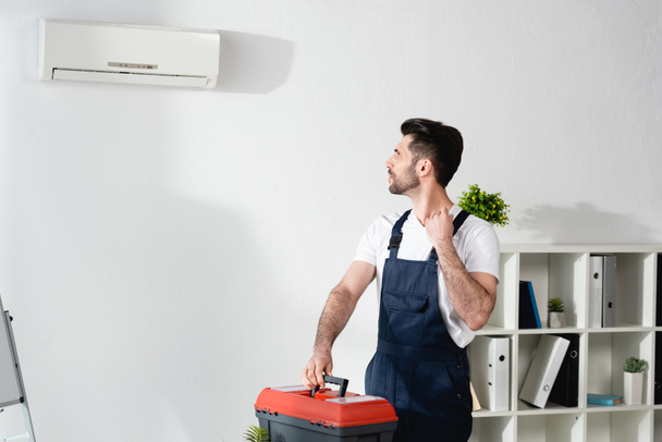 young repairman looking at air conditioner on wall while holding toolbox - Photo, Image