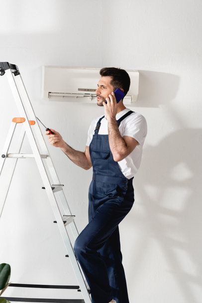 workman talking on smartphone and holding screwdriver on stepladder near air conditioner - Photo, Image