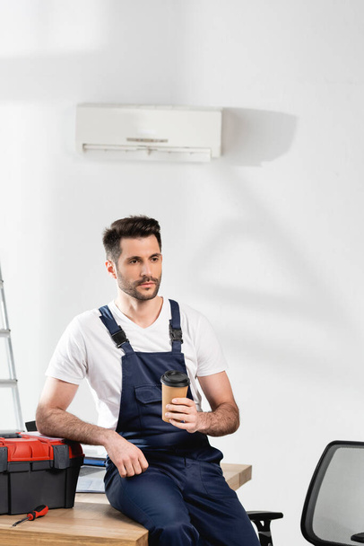 handsome repairman holding coffee to go while sitting at desk near toolbox under air conditioner on wall - Fotografie, Obrázek