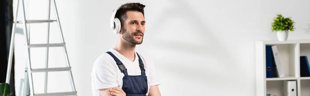horizontal image of repairman in wireless headphones standing with crossed arms near stepladder and air conditioner on wall - Photo, Image
