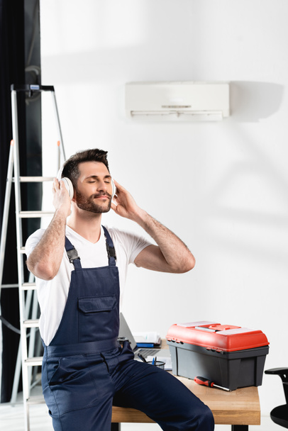 repairman with closed eyes sitting on desk in wireless headphones near toolbox and air conditioner on wall - Foto, afbeelding