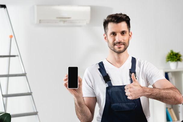 smiling repairman holding smartphone with blank screen and showing thumb up - Foto, imagen