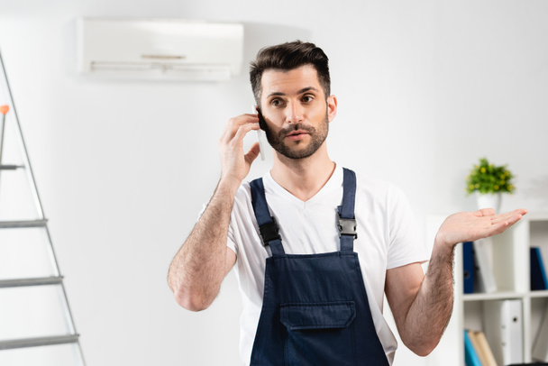 растерянный ремонтник разговаривает по смартфону и показывает жест пожатия плечами возле кондиционера на стене
 - Фото, изображение