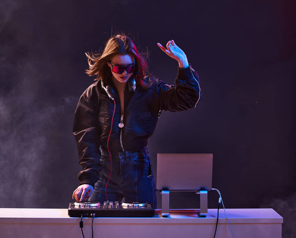 Stylish girl DJ in glasses headphones and a bomber mixes music dancing at a party in the neon light in the smoke . - Foto, Bild