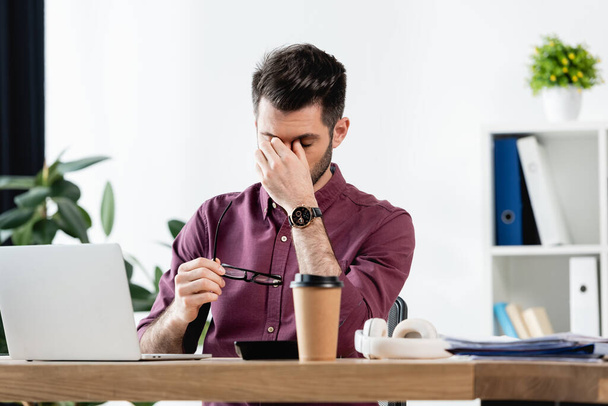 exhausted businessman holding eyeglasses and touching eyes while sitting near laptop  - Фото, изображение