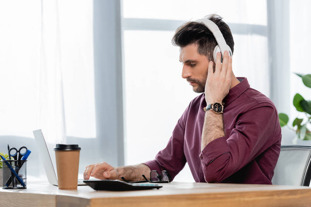 concentrated businessman in wireless headphones typing on laptop near coffee to go - Zdjęcie, obraz