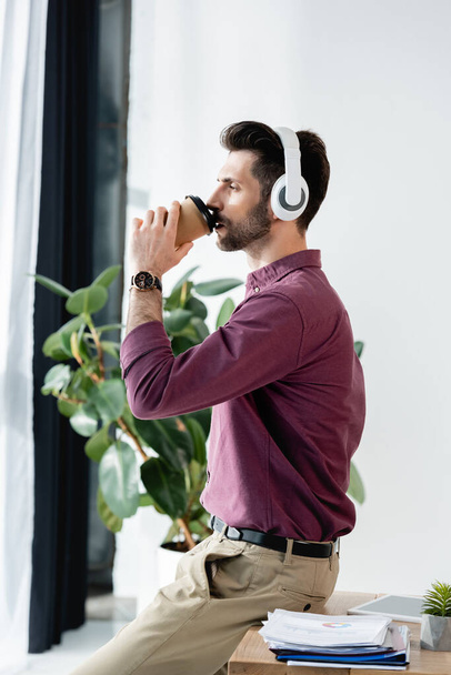 πλαϊνή άποψη του επιχειρηματία πίνοντας καφέ για να πάει, ενώ κάθεται στο γραφείο - Φωτογραφία, εικόνα