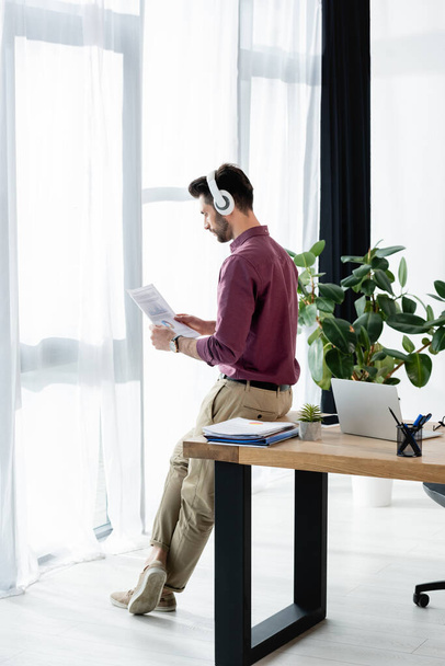 businessman in wireless headphones looking at document while sitting on desk near window - Fotoğraf, Görsel
