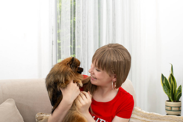 Постріл милої молодої жінки, що сидить на дивані і обіймає свого улюбленого пекінського собаку вдома. Любов між собакою і власником
. - Фото, зображення