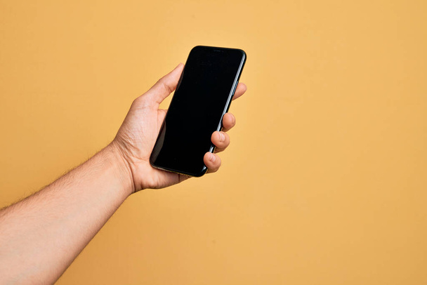 Hand of caucasian young man holding smartphone showing screen over isolated yellow background - Foto, Imagem