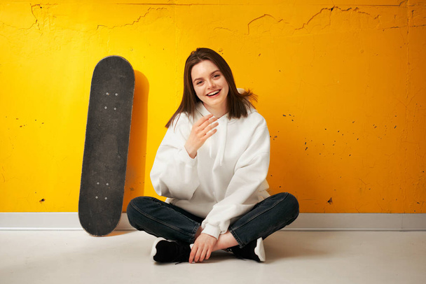 Young pretty happy smiling girl in a white hoodie with a skateboard poses against a yellow concrete wall. - Foto, imagen