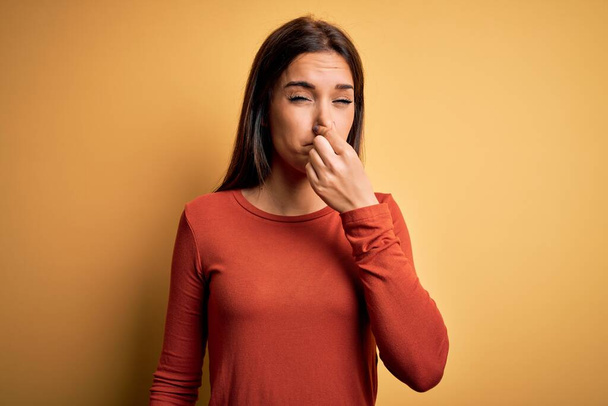 Young beautiful brunette woman wearing casual t-shirt standing over yellow background smelling something stinky and disgusting, intolerable smell, holding breath with fingers on nose. Bad smell - Photo, image