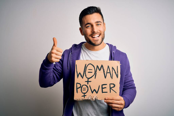 Young handsome activist man protesting for woman power holding cardboard banner happy with big smile doing ok sign, thumb up with fingers, excellent sign - Foto, immagini